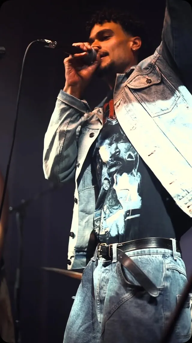 Osk on stage in a denim jacket over a vintage rap tee, singing into a mic under warm light.