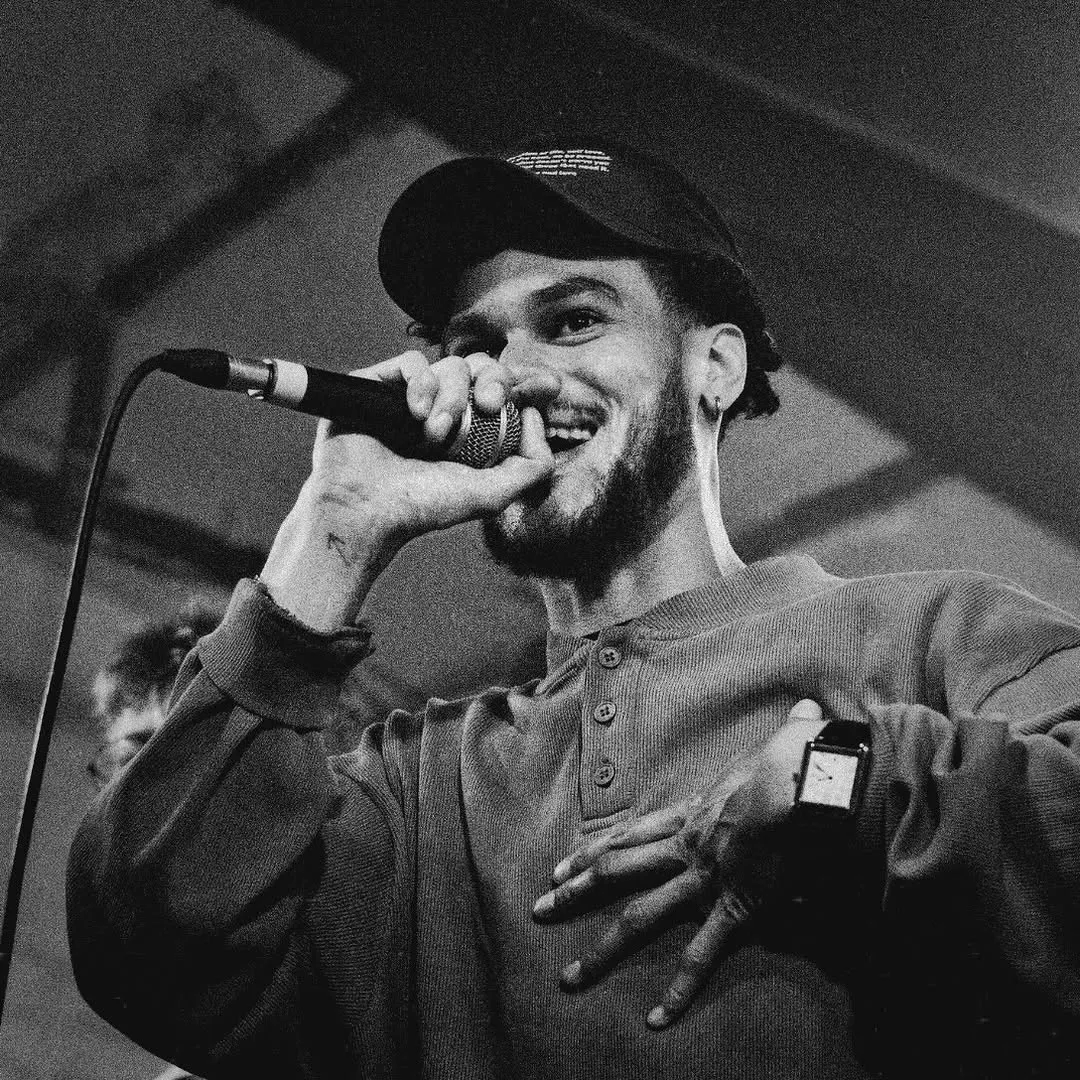 Black and white portrait of Osk in a cap, smiling at the mic with one hand on his chest.