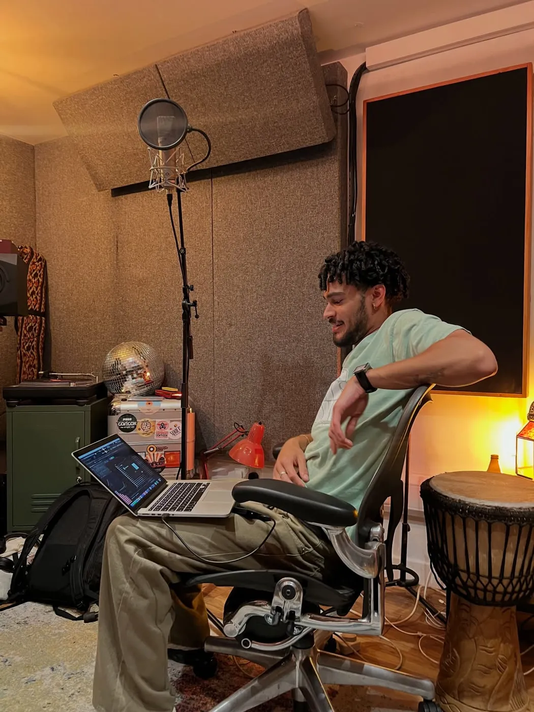 Osk leaning back in a desk chair in his home studio, laptop, microphone and a djembe beside him.