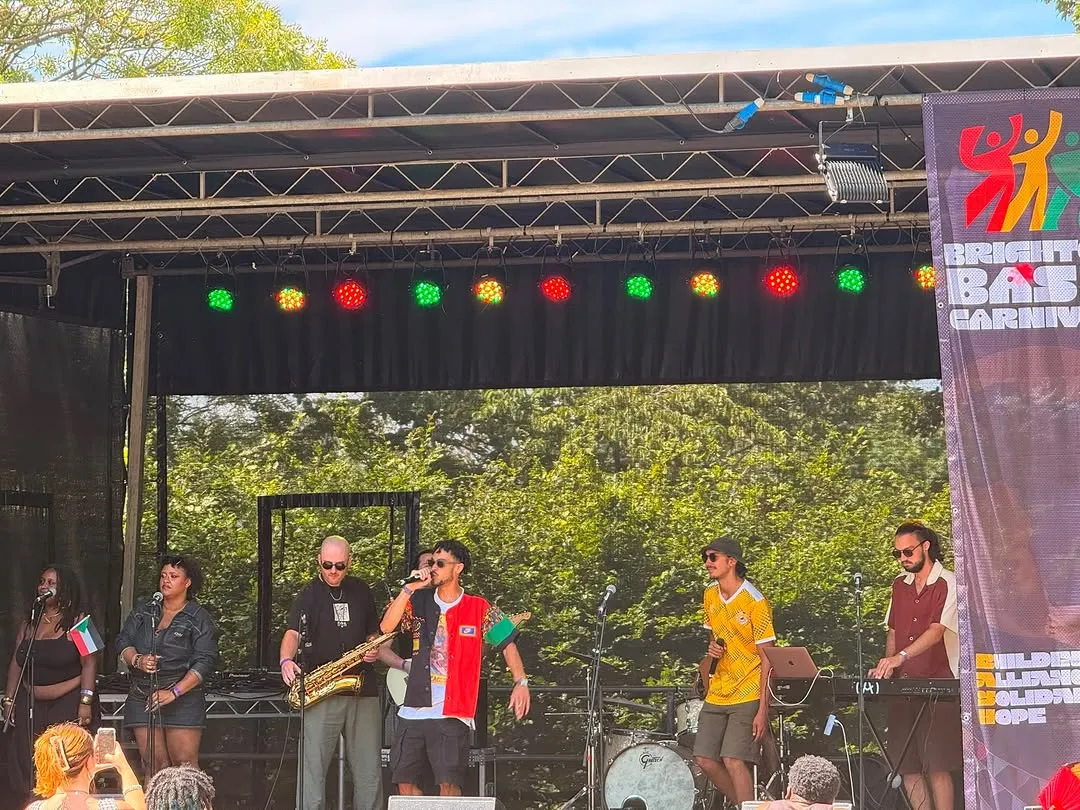Osk and his full band on an outdoor stage at BASH Carnival: saxophone, guitar and drums, trees behind and coloured lights above.
