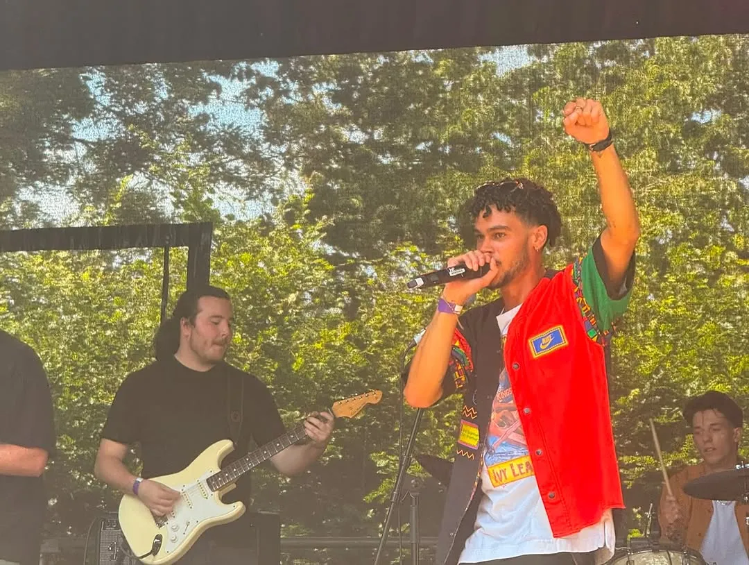 Osk with a raised fist, rapping into a mic on stage in a red jacket, guitarist behind him.