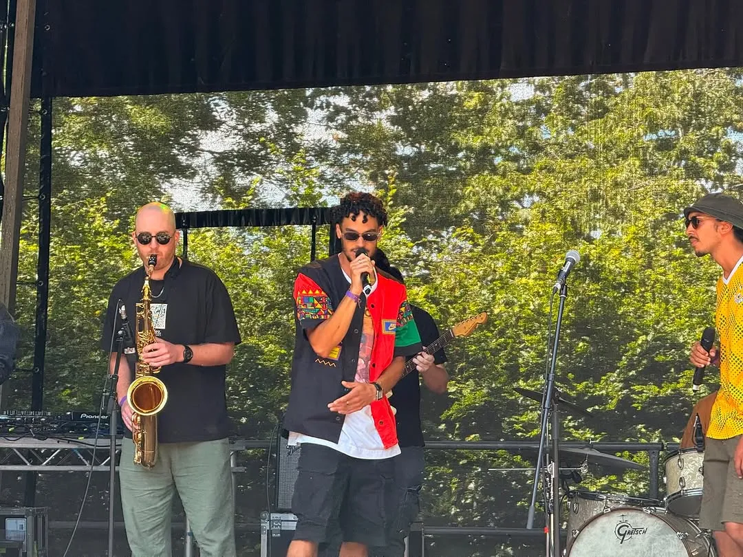 Osk in sunglasses and a red vest rapping beside the saxophonist on the outdoor stage.