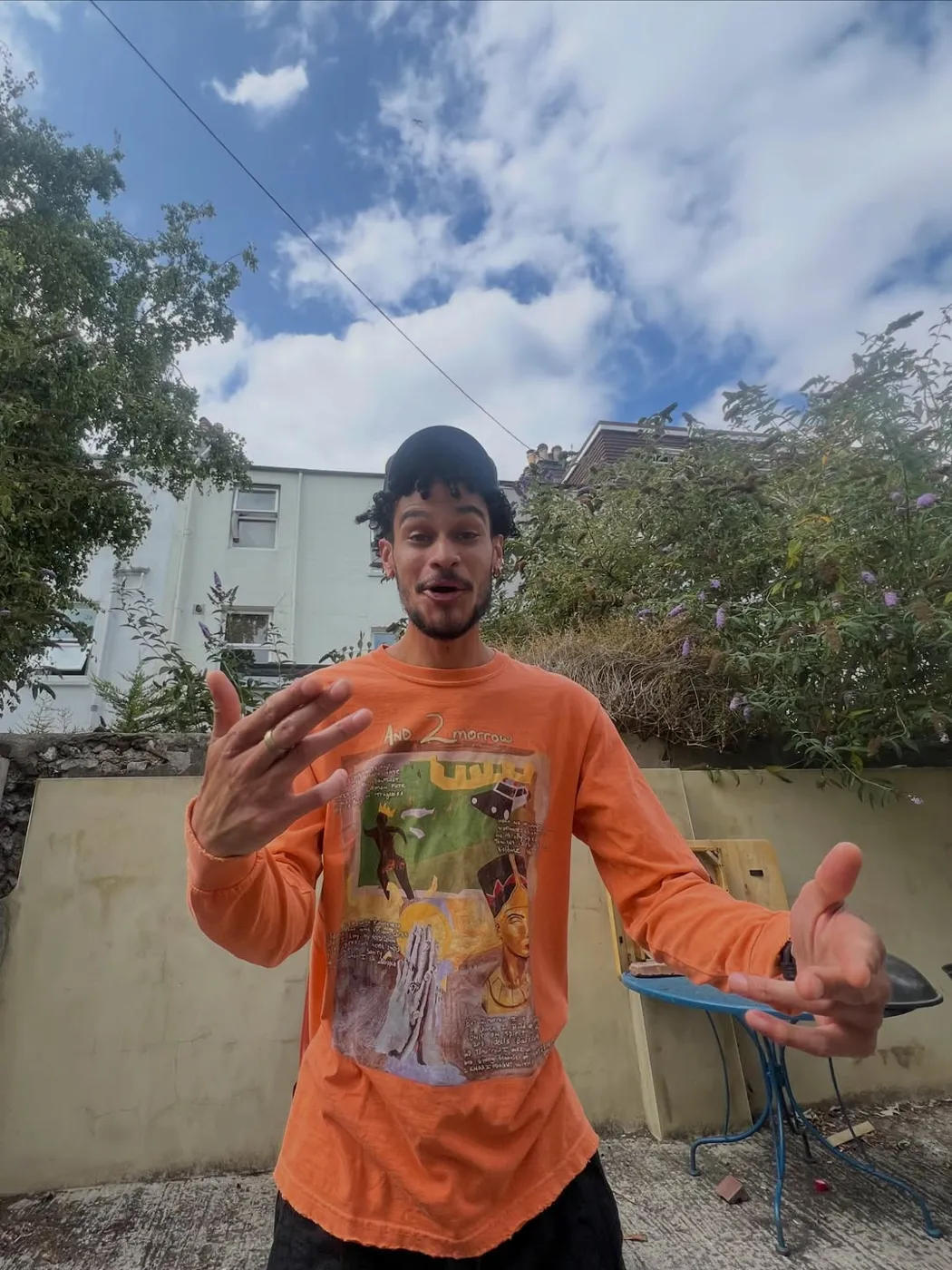 Osk in an orange graphic long-sleeve, mid-laugh with hands out, in a Brighton backyard.
